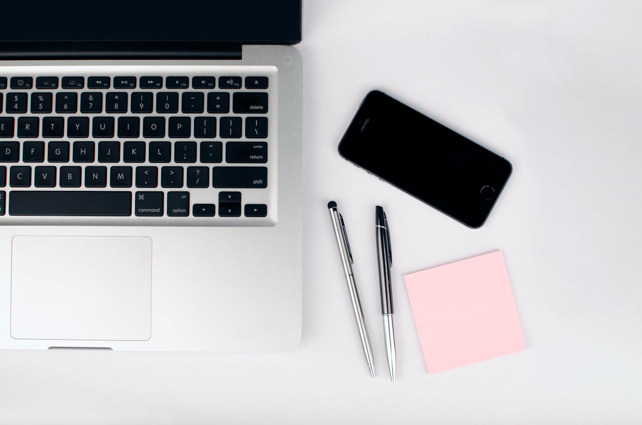 About A top view of a modern workspace featuring a laptop, smartphone, pens, and a pink sticky note on a white desk.